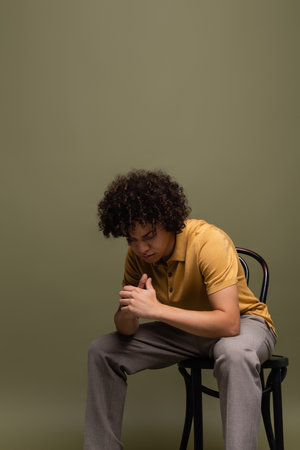 curly african american man in yellow polo shirt and trousers sitting on chair with bowed head on grey green backgroundの写真素材