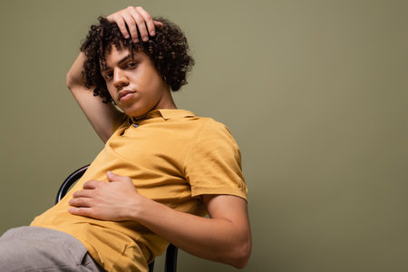 curly and stylish african american man in yellow polo shirt sitting with hand on head isolated on greyの写真素材