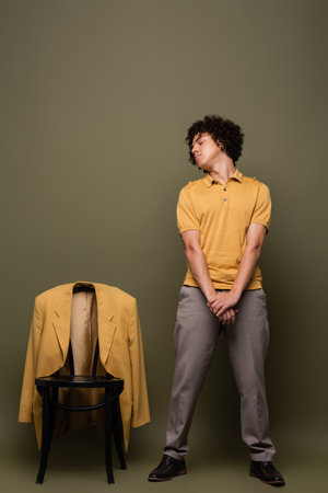 full length of african american man in yellow polo shirt and pants standing with closed eyes near chair with blazer on grey green backgroundの写真素材