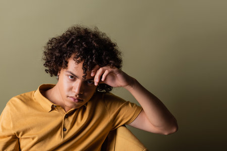 curly african american man in yellow polo shirt holding hand near head and looking at camera while sitting on grey green backgroundの写真素材