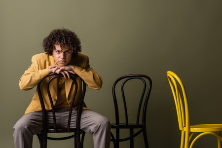 brunette african american man in trendy casual outfit sitting on black chair and looking at camera on olive grey backgroundの写真素材