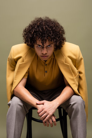 young and confident african american man in yellow jacket looking at camera while sitting isolated on greyの写真素材