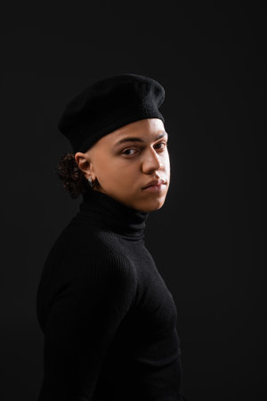 portrait of young african american man in trendy beret and turtleneck looking at camera isolated on blackの写真素材