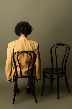 back view of brunette african american man with curly hair sitting in yellow blazer on black chair on grey green backgroundの写真素材