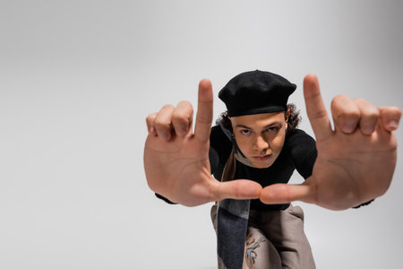 african american man in black beret and warm scarf looking at camera and showing snapshot gesture on greyの写真素材
