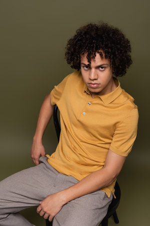 young african american man in yellow polo shirt sitting on chair and looking at camera on grey green backgroundの写真素材
