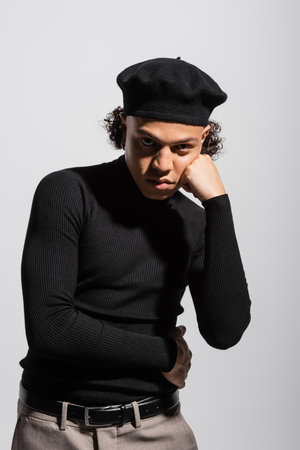 young and trendy african american man in black beret and turtleneck standing with hand near face and looking at camera isolated on greyの写真素材