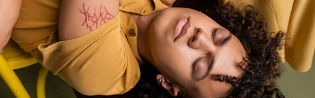 top view of tattooed african american man with closed eyes lying in yellow polo shirt on grey green background, bannerの写真素材