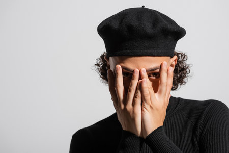 young african american man in black stylish beret obscuring face with hands and looking at camera isolated on greyの写真素材