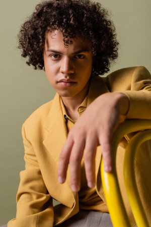 stylish african american man in yellow jacket sitting and looking at camera on blurred foreground isolated on greyの写真素材