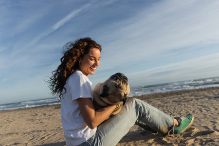 happy young woman with curly hair holding pug dog while sitting on beach near sea in Spainの写真素材