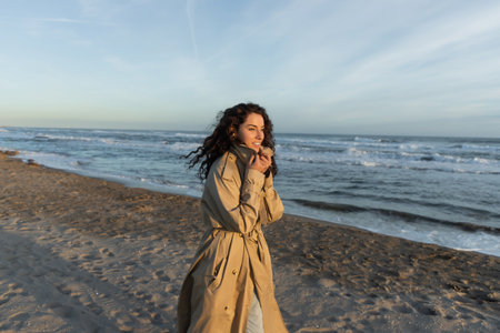 cheerful woman in stylish beige trench coat walking on beach in Barcelonaの写真素材