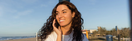 cheerful young woman talking on smartphone on beach in Barcelona, banner