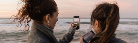 Friends taking photo on smartphones near sea during sunset, bannerの写真素材