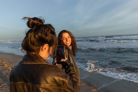 Woman taking photo on smartphone of smiling friend on beach in Barcelonaの写真素材