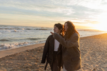 Positive woman pointing with hand while talking to friend on beach near sea