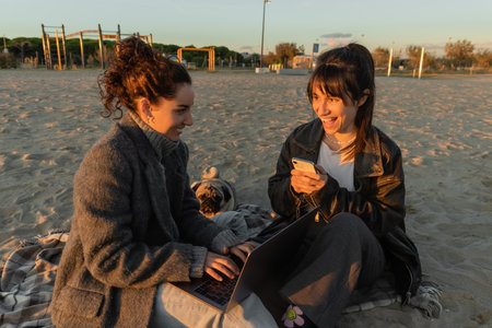 Cheerful women using gadgets near pug dog on beach in Spainの写真素材