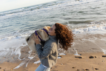 happy and curly young woman holding pug dog on beach near sea in Barcelonaの写真素材