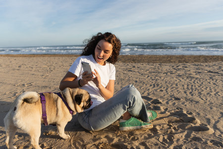 cheerful woman in white t-shirt taking photo of pug dog on sandy beach in Barcelonaの写真素材