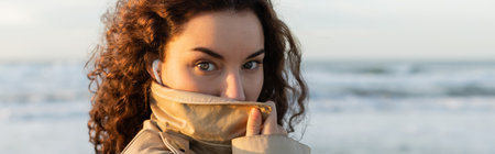 portrait of curly woman in wired earphones adjusting collar of beige trench coat and looking at camera near sea, bannerの写真素材