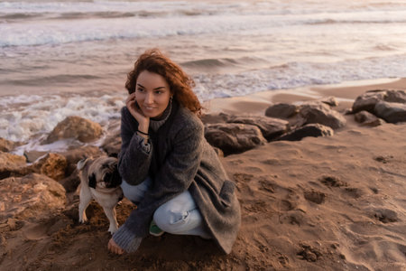Smiling woman in coat looking away near pug dog on beach in Barcelonaの写真素材