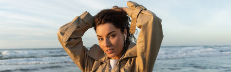 young woman in beige trench coat and wired earphones listening music while adjusting hair near sea...