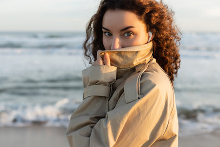 portrait of curly woman in wired earphones adjusting collar of beige trench coat and looking at...