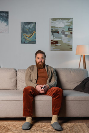Bearded man in cardigan holding smartphone while sitting on couch at homeの写真素材