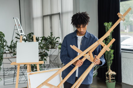 Young african american artist holding wooden easel in workshop