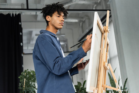 African american artist painting on canvas on easel in studio