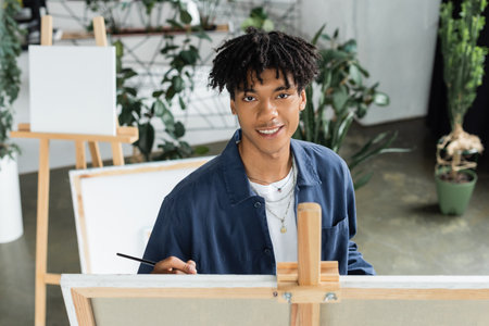 Smiling african american artist holding paintbrush and looking at camera near canvas in studio