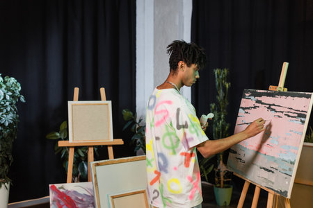 Side view of african american artist painting on canvas near drawings in studio