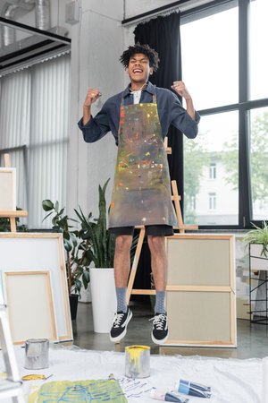 Excited african american artist in apron jumping in studio