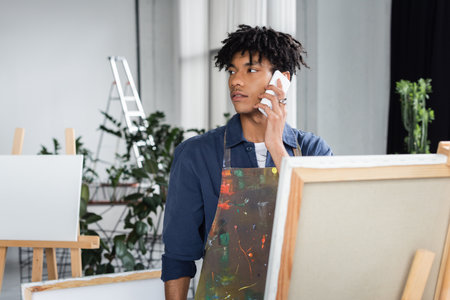 Young african american artist in apron talking on cellphone in workshopの写真素材