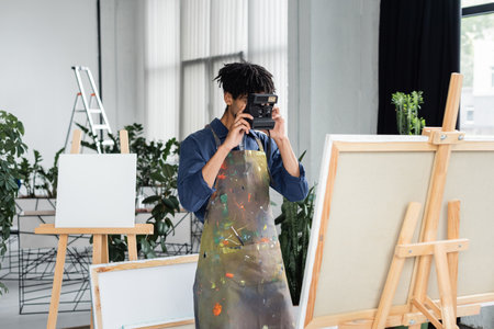Young african american artist in apron taking photo on vintage camera near canvas in studio