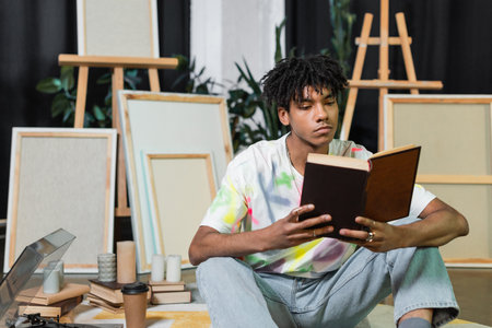 African american artist reading book near record player and canvases in workshop