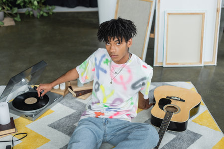 Young african american artist using record player near acoustic guitar and canvases in studio