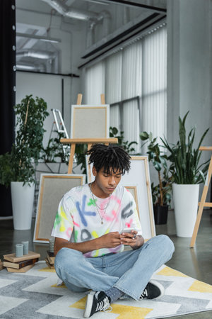 Young african american artist using smartphone near books and canvases in art studioの写真素材