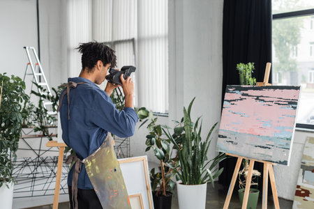 African american artist taking photo on vintage camera near painting in studio