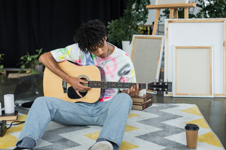 African american artist playing acoustic guitar near canvases and coffee in studio