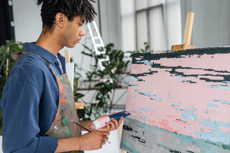 Side view of african american artist holding paint and paintbrush near abstract painting in studio