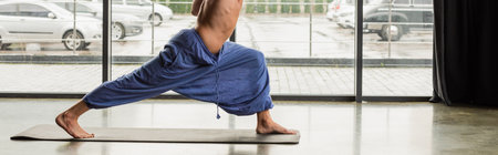 cropped view of shirtless man in pants doing crescent lunge pose on yoga mat, banner