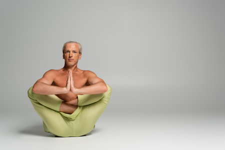 shirtless man in pants sitting in womb embryo pose and doing anjali mudra on greyの写真素材