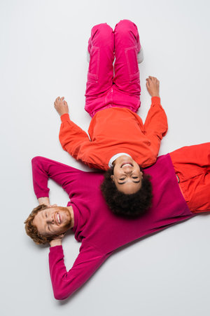 top view of cheerful redhead man and happy african american woman in magenta color clothes lying on...