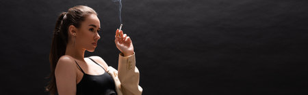 young brunette woman in beige blazer holding cigarette while looking away on black background, bannerの写真素材