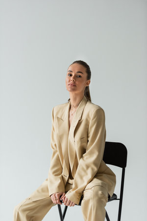 brunette young woman in beige trendy suit sitting on chair isolated on greyの写真素材