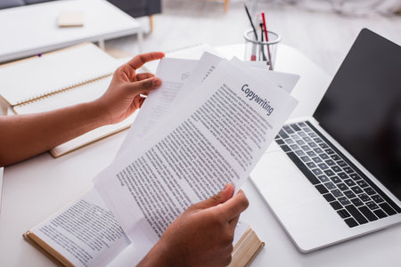 Cropped view of freelancer holding papers with copywriting lettering near book and laptop at home