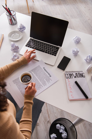 Top view of copywriter holding coffee and using laptop near crumpled paper at homeの写真素材
