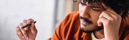 pensive indian man holding pen while looking away at home, banner