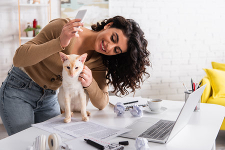 Cheerful copywriter holding smartphone and petting oriental cat near papers and laptopの写真素材